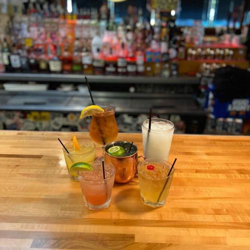 A variety of colorful cocktails on a wooden bar, with a blurred bar background.