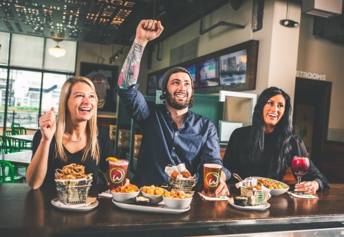 Three friends celebrate at a restaurant, enjoying food and drinks, with one person raising a fist in excitement.