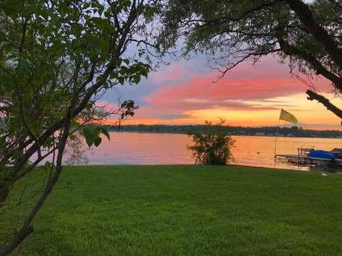 A serene lakeside view at sunset, with vibrant pink and orange skies, green grass, and a flag by the water.