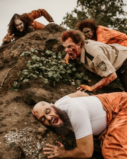A group of four people in zombie costumes, covered in dirt and fake blood, climbing a mound of earth in a spooky setting.