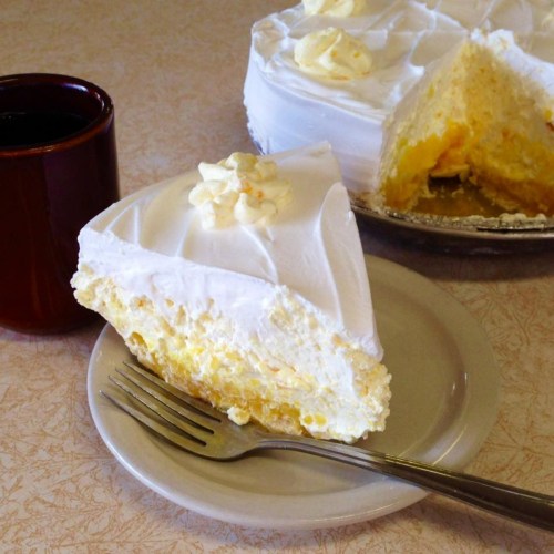A slice of creamy lemon pie with whipped topping on a plate, next to a cup of coffee.