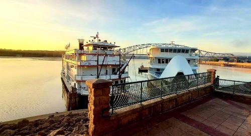 Two riverboats docked at a serene waterfront during sunrise, with a bridge in the background and calm waters.