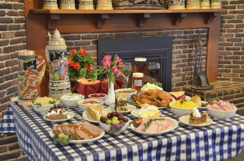 A festive table spread with various dishes, drinks, and desserts, set against a rustic brick fireplace backdrop.