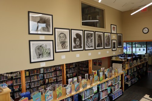 A library interior featuring a wall of framed artwork above shelves filled with children's books.