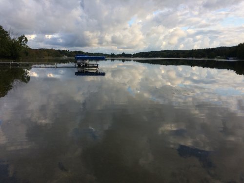 A calm lake reflecting clouds and trees, with a small dock visible on the water's surface.
