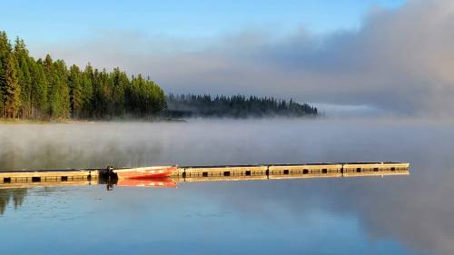 A serene lake scene with a foggy shoreline, a wooden dock, and lush green trees in the background.