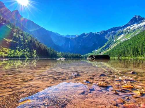 A serene mountain lake reflecting blue skies and sunlight, surrounded by lush greenery and rocky peaks.