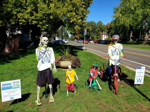 Three skeletons in sports attire stand on a lawn, promoting walking and biking, with signs nearby.