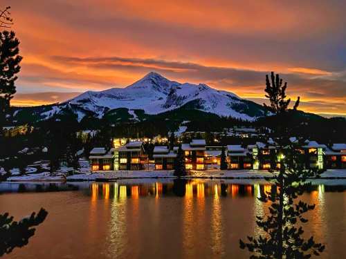 A scenic winter sunset over a snowy mountain, reflecting on a calm lake with cozy cabins illuminated along the shore.