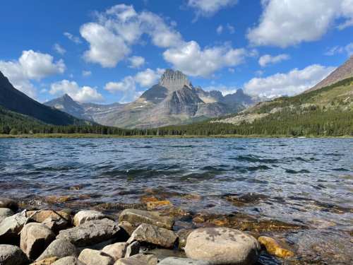A serene lake surrounded by mountains under a blue sky with fluffy clouds, featuring a rocky shoreline in the foreground.