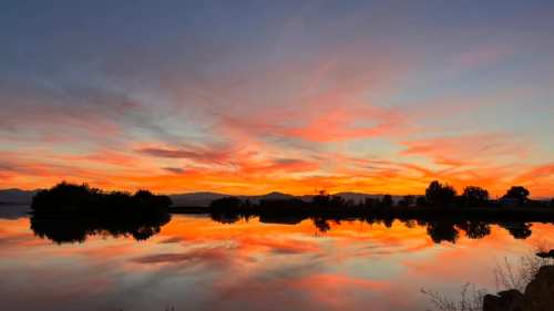 A vibrant sunset reflects on a calm lake, with colorful clouds and silhouettes of trees in the foreground.