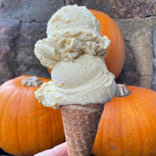 A hand holding a cone with two scoops of creamy ice cream, set against a backdrop of orange pumpkins.