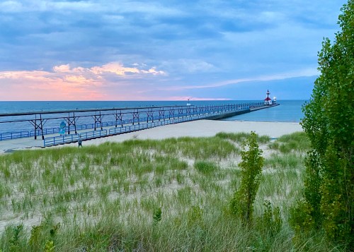 A serene beach scene with a pier extending into the water, surrounded by sand dunes and greenery under a cloudy sky.