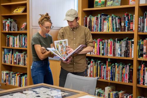 A woman and a man examine a book together in a library filled with shelves of colorful books.