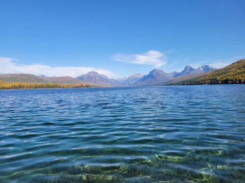 A serene lake with clear water, surrounded by mountains and a blue sky, reflecting a peaceful natural landscape.