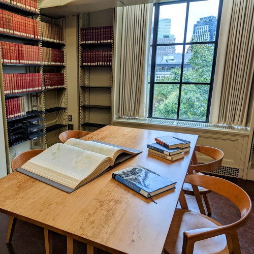 A spacious library table with an open book and several stacked books, surrounded by shelves and a large window view.