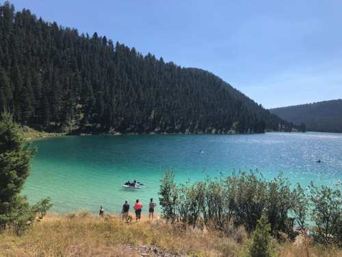 A serene lake surrounded by green trees, with people enjoying the water and a boat in the distance under a clear blue sky.