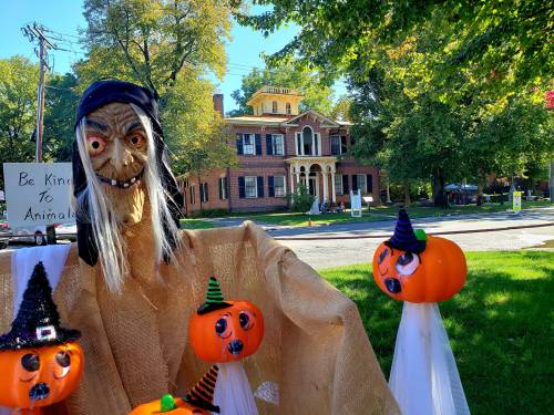 A spooky scarecrow with pumpkins in front of a large, historic house, surrounded by autumn trees.