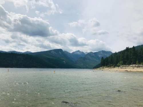 A serene lake surrounded by mountains and trees, with a cloudy sky above and a few people enjoying the water.