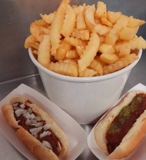 A bowl of crinkle-cut fries next to two hot dogs topped with onions and green relish.
