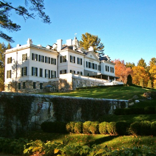 A large, elegant mansion with white walls and black shutters, surrounded by lush greenery and colorful autumn trees.