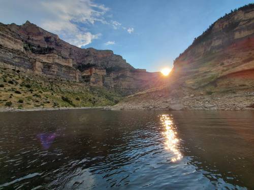 Sunset over a serene river, reflecting light on the water, surrounded by towering cliffs and lush greenery.
