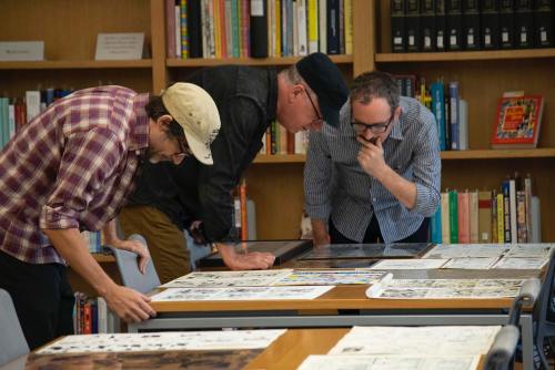 Three men examine documents on a table in a library, surrounded by bookshelves filled with books.