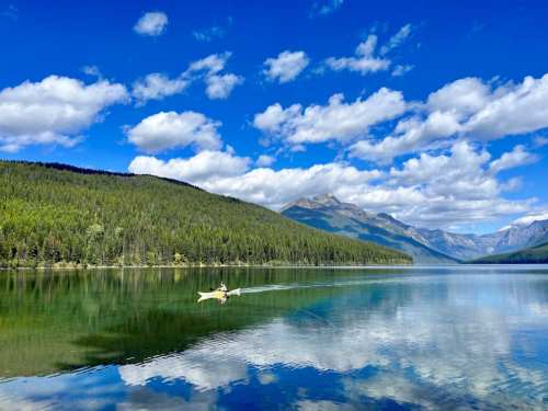 A serene lake reflects mountains and clouds, with a person kayaking in the foreground surrounded by lush greenery.