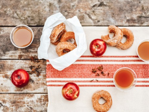 A rustic table with donuts, red apples, and cups of cider on a striped cloth.