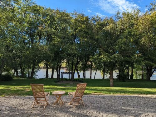Two wooden chairs and a small table facing a serene river view, surrounded by lush green trees under a blue sky.