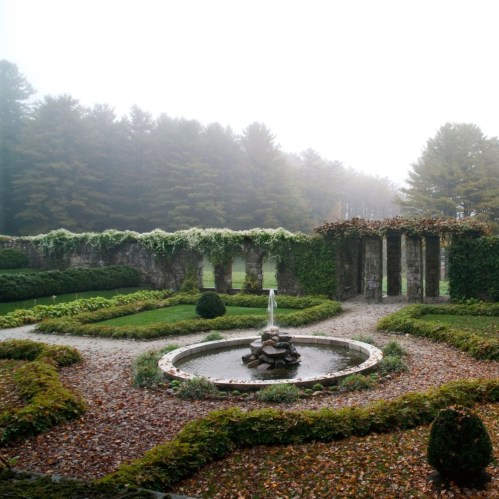 A misty garden with a circular fountain surrounded by stone columns and lush greenery.