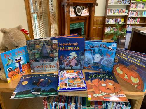 A table in a library displaying various children's books with Halloween themes, surrounded by a cozy atmosphere.