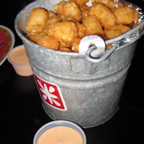 A metal bucket filled with golden, crispy tater tots, accompanied by two small cups of dipping sauce.