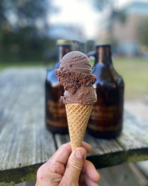 A hand holding a chocolate ice cream cone with two scoops, set against blurred bottles in the background.