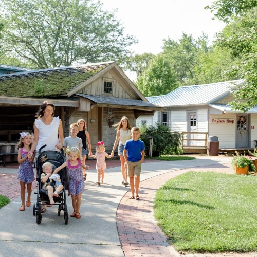 A woman pushes a stroller while walking with several children along a path in a sunny outdoor setting.