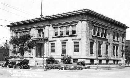 Historic black-and-white photo of a large, classical-style library building with a vintage car parked in front.