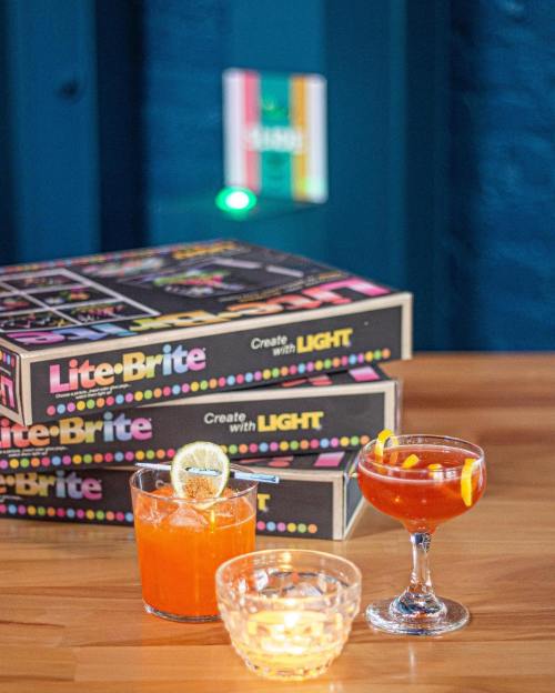A colorful display featuring Lite-Brite boxes and two cocktails on a wooden table, with a glowing light in the background.