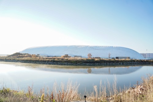 A large, white dome structure reflected in a calm body of water, surrounded by grass and trees under a clear sky.