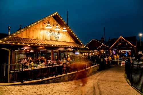 A lively bar with outdoor seating, illuminated by string lights, bustling with people at dusk.