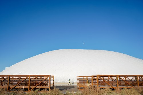 A person walks near a large, white dome structure under a clear blue sky with a small moon visible.