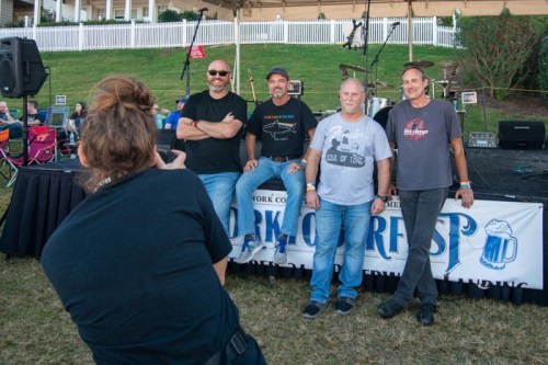 A photographer captures four men posing on stage at an outdoor event, with a grassy area and equipment in the background.
