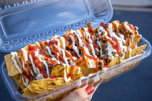 A hand holding a clear container filled with nachos topped with meat, mayonnaise, and ketchup drizzles.
