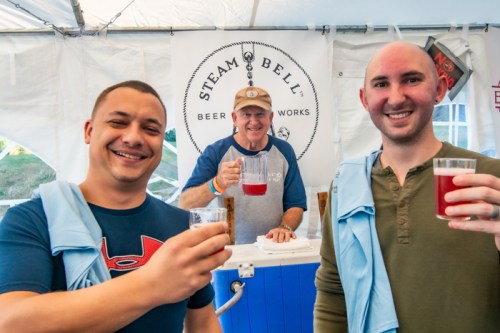 Three men smile and raise glasses of beer in a tent, with a sign for Steam Bell Beer Works in the background.