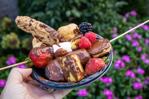 A hand holding a plate of chocolate-covered strawberries, marshmallows, and cookies, surrounded by colorful flowers.