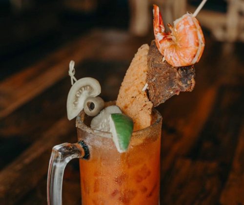 A tall glass of a colorful cocktail garnished with shrimp, lime, and a fried snack, resting on a wooden table.