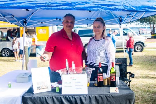 A man and woman stand behind a table at a market, showcasing wine bottles under a blue tent.