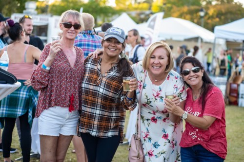 Four women smile and pose together at an outdoor event, holding drinks, with festival tents in the background.