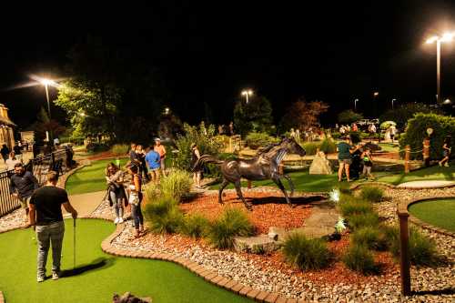 A lively mini-golf course at night, featuring a horse statue and groups of people enjoying the game.