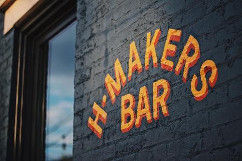 Colorful sign reading "H-MAKERS BAR" on a textured brick wall, with a window reflecting the sky.