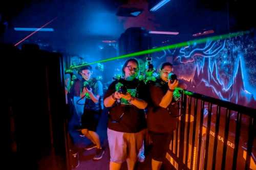 A group of people in a dark, neon-lit room, aiming laser tag guns with colorful laser beams in the background.